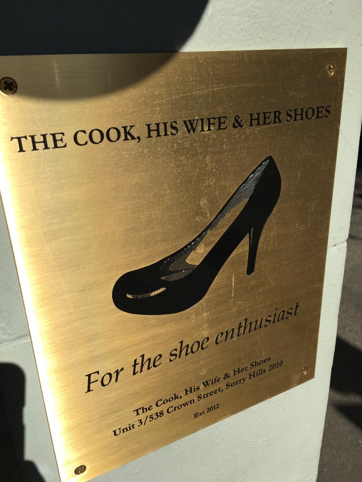 A Gold Sign Outside of The Cook, His Wife & Her Shoes Shop that Says, "For the Shoe Enthusiast."