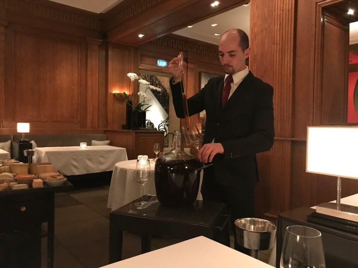 A photo of a sommelier serving port wine from the French side of southwest Andorra being served out of a giant glass jug and syringe at Le Taillevent in Paris, France.