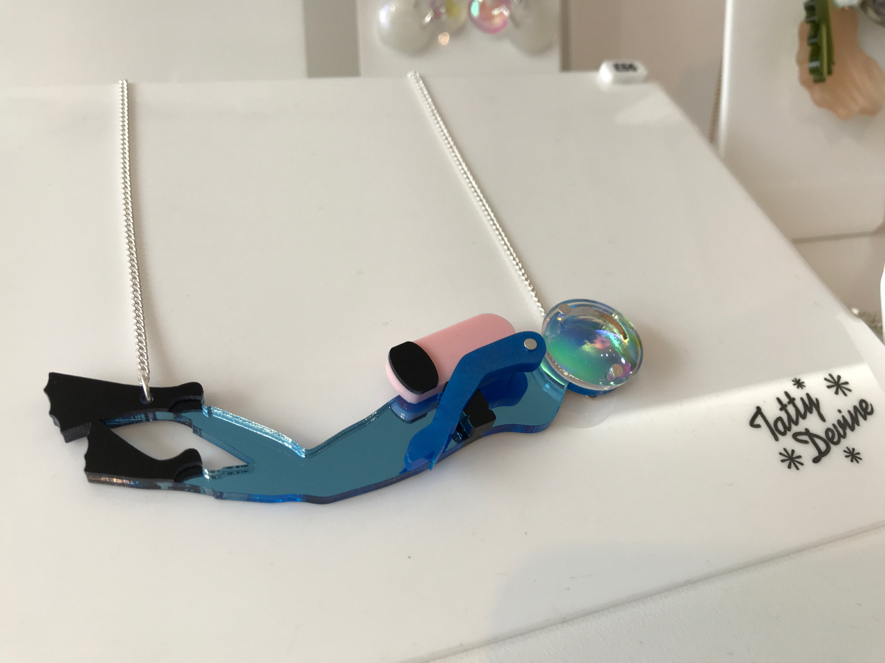 A photo of the, "Diver Necklace," from Tatty Devine. Tatty Devine is a local shop located in London, England. The necklace features a blue female scuba diver with a pink scuba tank made out of mirror and pearlescent acrylic. The necklace hangs from a silver necklace chain.