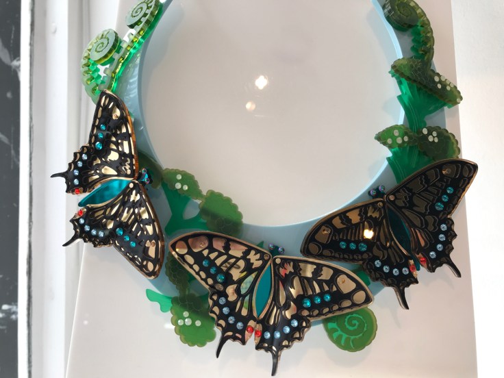 A photo of the limited edition, "Swallowtail Statement Necklace," from Tatty Devine. Tatty Devine is a local shop located in London, England. The necklace features iridescent butterflies in gold, blue and orange surrounded by green ferns. The butterflies are made out of Swarovski crystals and mirror acrylic. The necklace is in a collar shape.