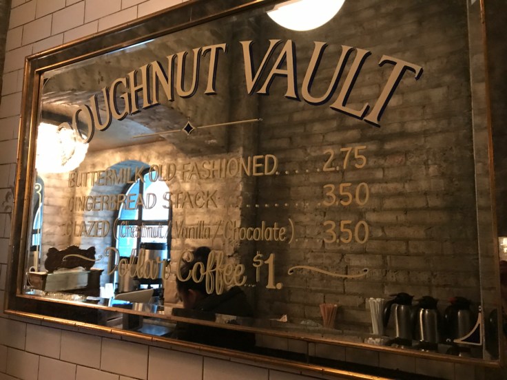 A photo of a mirrored sign at the Doughnut Vault in Chicago, Illinois. The sign says, "Doughnut Vault Buttermilk Old Fashioned $2.75 Gingerbread Stack $3.50 Glazed (Chestnut / Vanilla / Chocolate) $3.50 Dollar Coffee $1."