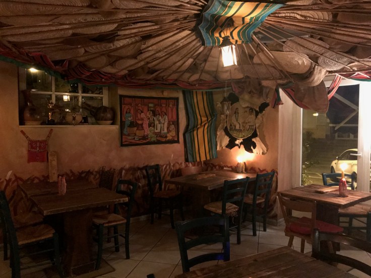 A photo of the inside dining room at Ambiente Africa in Stuttgart, Germany. There are colorful cushions and mats as well as embroidered goat skins and hand carved sculptures inside the restaurant.