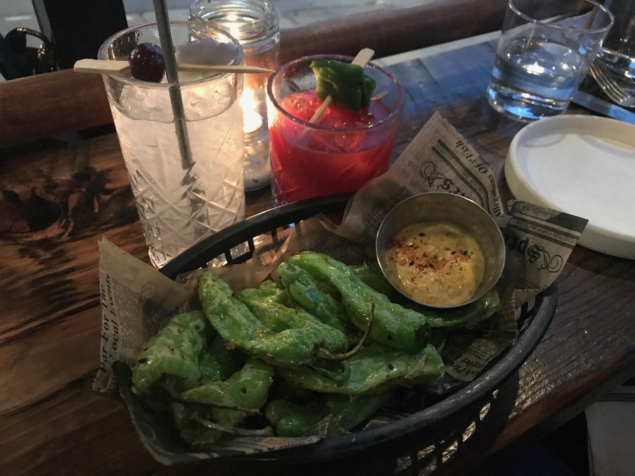 A photo of the aviation cocktail which is served on tap at The Snug SF restaurant in San Francisco, California. The clear cocktail is made with crème de violette, maraschino, sparkling and lemon. Beside it sits the red hued prickly pear. The prickly pear is made with mezcal, amaro, nopales, prickly pear, habanero and lime. In front of the two drinks sit a basket filled with shishito peppers and sweet corn togarashi dip.