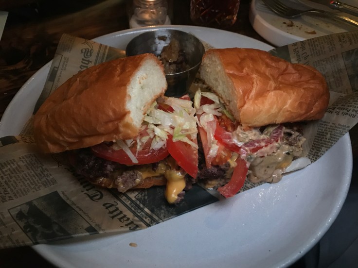 A photo of the bodega "burger" at The Snug SF in San Francisco, California. The burger comes with Wagyu beef, lettuce, tomato, onion, cheese and a special sauce.
