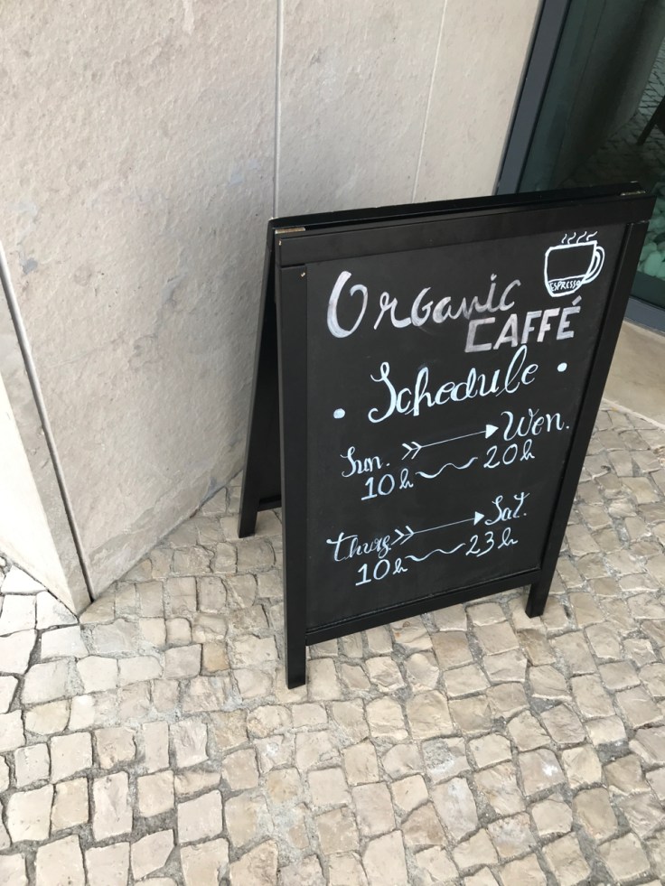 A photo of a sign outside the exterior of Organic Caffe in Estoril, Portugal. The sign has the schedule for the cafe written on it.