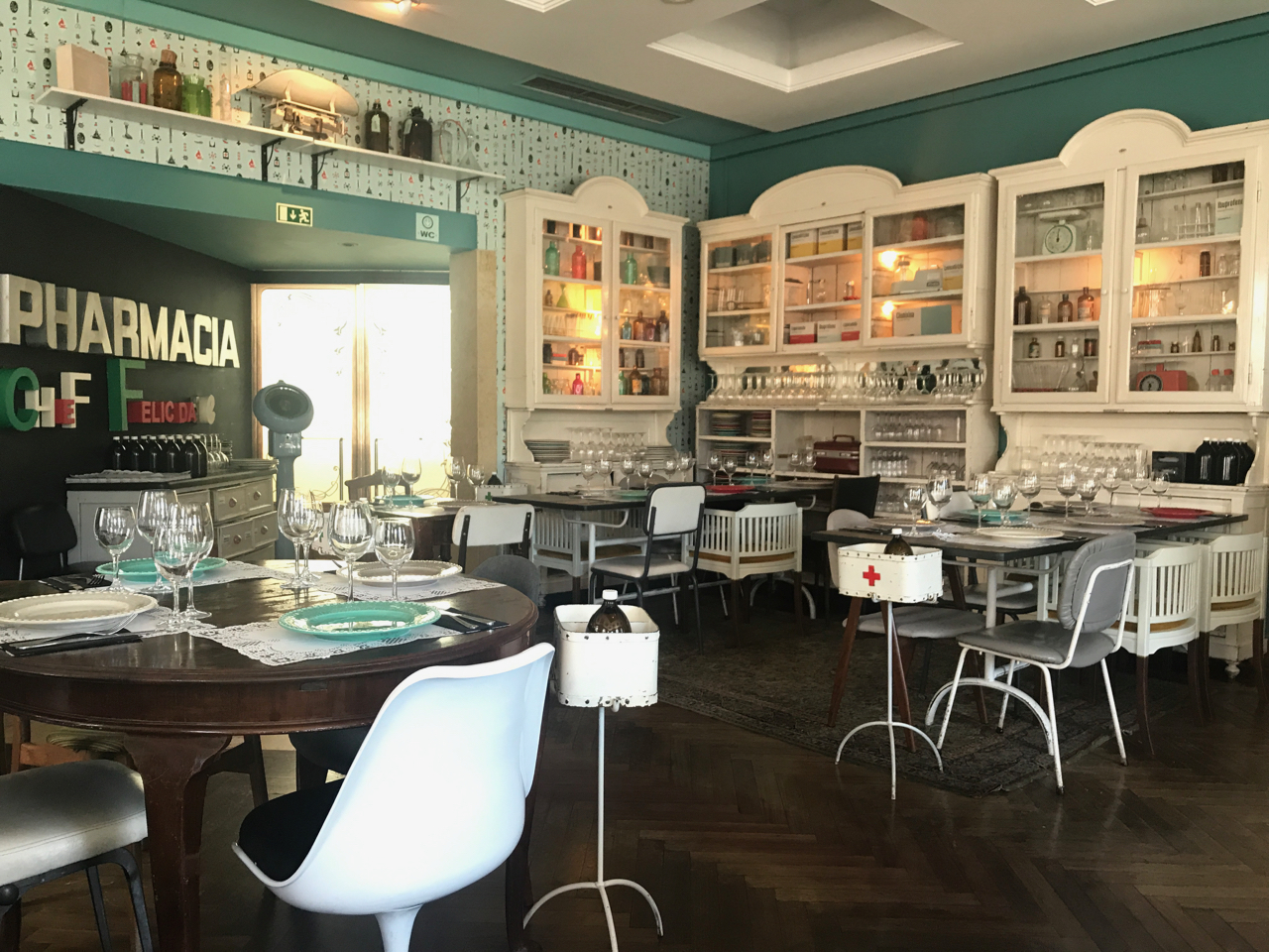 A photo of the tables, chairs and wallpaper (all of which are pharmacy themed) at Pharmacia restaurant in Lisbon, Portugal. All of the shelves and cabinets are filled with old drug bottles and other objects you might find at a pharmacy.