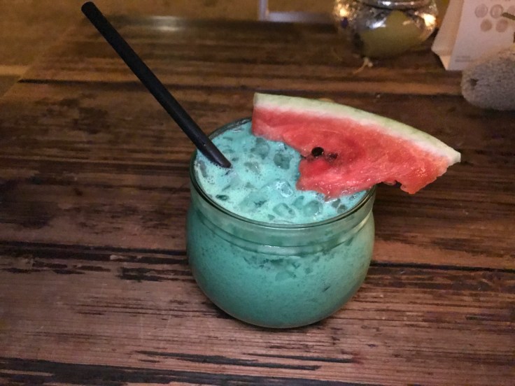 A photo of a bright blue cocktail topped with a slice of watermelon that is called the Kilimanjaro at Ambiente Africa restaurant in Stuttgart, Germany. The Kilimanjaro is made with amarula cream liqueur, orange and banana juice. (Kilimanjaro - Amarula, orange und bananensaft)