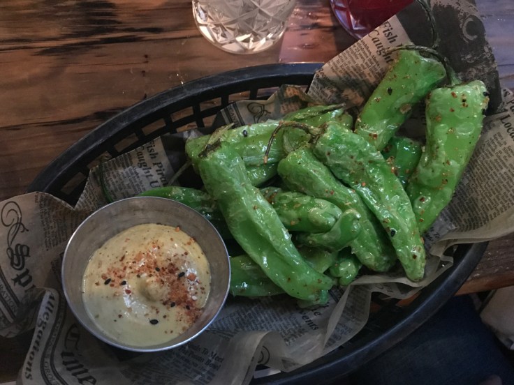 A photo of a black basket filled with green steamed shishito peppers served with sweet corn togarashi dip at The Snug SF in San Francisco, California.