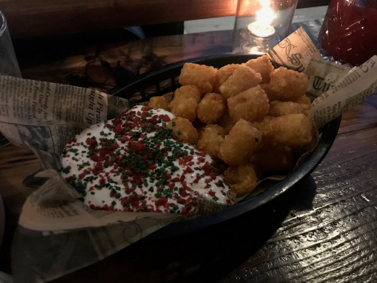 A photo of a black basket filled with tater tots at The Snug SF in San Francisco, California. On the side is a horseradish sour cream topped with chives and Bac-Os.