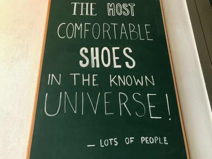 A photo of a chalkboard sign that says, "The most comfortable shoes in the known universe!" The bottom of the sign says according to, "Lots of People," spotted at Atheist Shoes in Berlin, Germany.