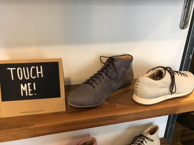 A photo of a kitten testicle grey boot and a Nabakov cream sneaker on a wood shelf beside a sign that says, "Touch me!" at Atheist Shoes in Berlin, Germany.