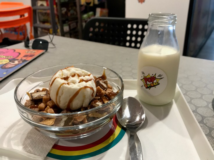 A cereal bowl and a jug of milk sit on a cafeteria style lunch tray at Pop Cereal Café in Porto, Portugal. The item on the tray is the, "#3. My Friends Call Me Caramel." It's Made With Nestlé Lion Cereal, Golden Grahams, Cookie Crisp, Caramel Chocolate, Toffee, Toffee Topping and Milk