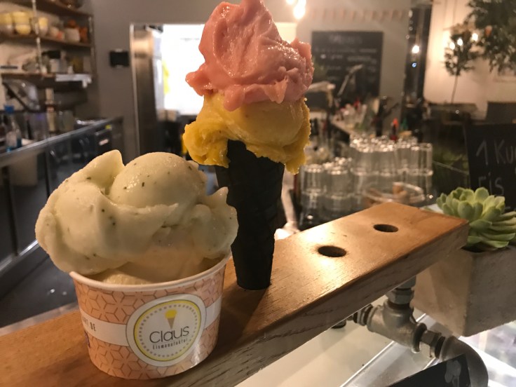 A photo of a cup of ice cream with scoops of buttermilk passionfruit (buttermilch maracuja) and basil smash made with lemon (zitrone basilikum) and a cone topped with vegan watermelon (wassermelone) and mango chili at Claus Eismanufaktur & Deli (gelato, ice cream and sorbet) in Stuttgart, Germany.