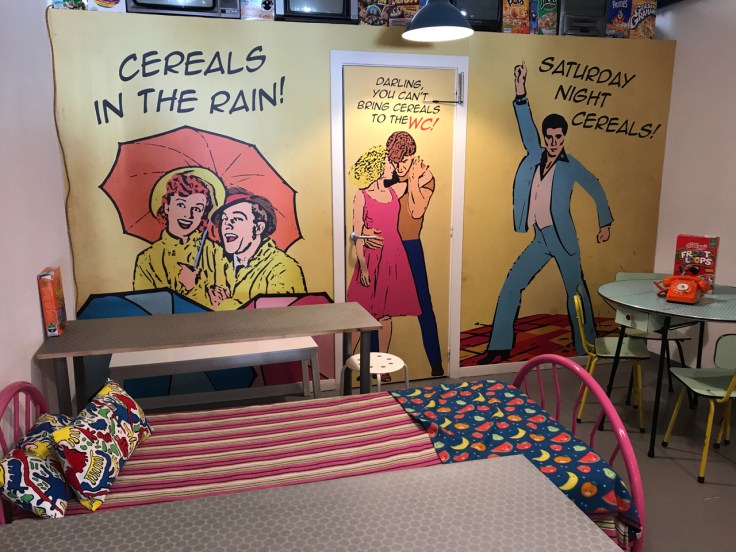 Graphics on the wall at Pop Cereal Café in Porto, Portugal say, "Cereals in the Rain," "Darling, You Can't Bring Cereals to the WC!" and "Saturday Night Cereals!" Seating options include beds as well as traditional restaurant tables and chairs.