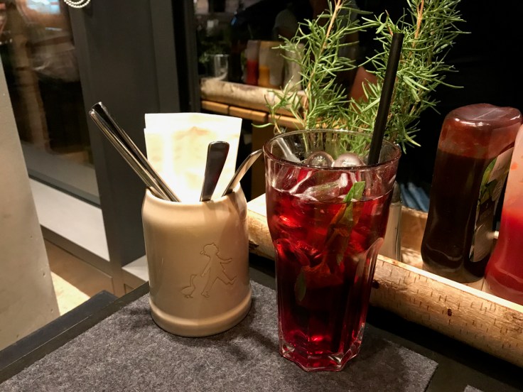 Beside a cure jar with a boy and a duck sits a johannisbeer basilikum (moranga lemonade with basil, currant and elderflower) at Hans im Glück burger grill in Stuttgart, Germany. The ruby red beverage sits is in front of a live little rosemary plant on the restaurant's table. Photo Courtesy of FoodWaterShoes