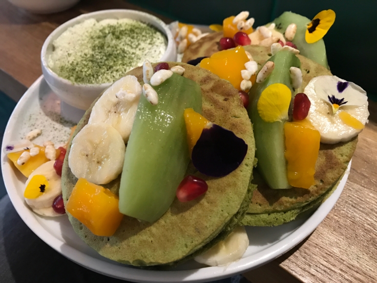 A plate is crammed full of three organic matcha pancakes (pancakes au matcha bio) at Peonies café and flower shop in Paris, France. The green pancakes are topped with flower petals, slices of banana, kiwi, pomegranate seeds and other brightly colored kinds of fresh seasonal fruit.