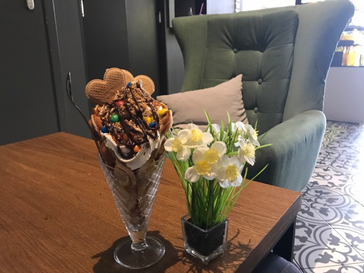 Zachcianka - A bubble waffle with whipped cream, two scoops of ice cream, M&M's, chocolate glaze and crushed peanuts sits beside a vase of flowers on a table at Ciekawa Słodko-Słono restaurant and ice cream shop in Tarnów, Poland.