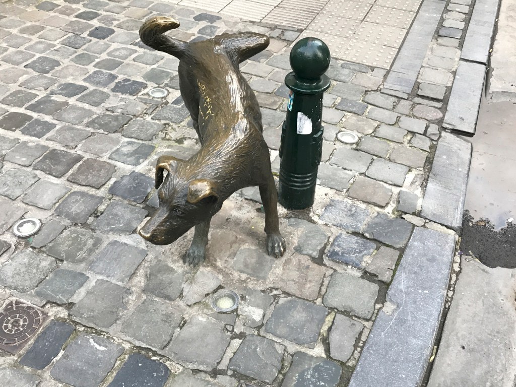 A photo of Zinneke Pis, a bronze statue of a dog peeing on a street pole in Brussels (or Bruxelles), Belgium. The statue is one of the lesser known landmarks you can check out at the corner of Rue des Chartreux and Rue du Vieux Marché aux Grains. Photo Courtesy of FoodWaterShoes.