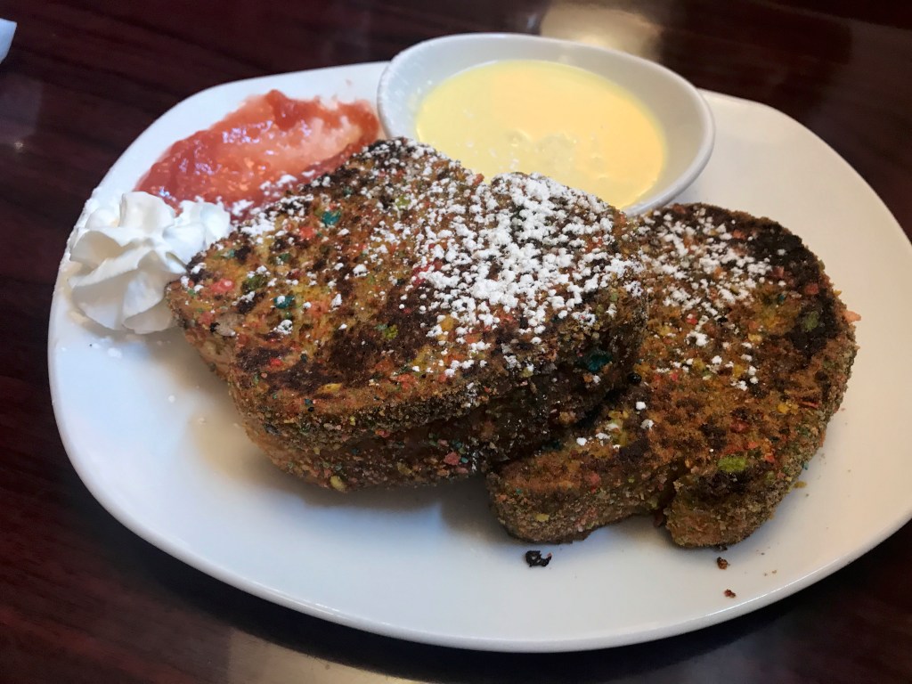 A photo of the Barney Rubble at Modern Malt in Syracuse, New York, The dish features two slices of French toast encrusted with rainbow hued Fruity Pebbles cereal. On the side there is red strawberry ginger jam and also a dish of crème anglaise for dipping. The quirky creative brekkie breakfast food looks weird, but tastes delicious. Photo Courtesy of FoodWaterShoes