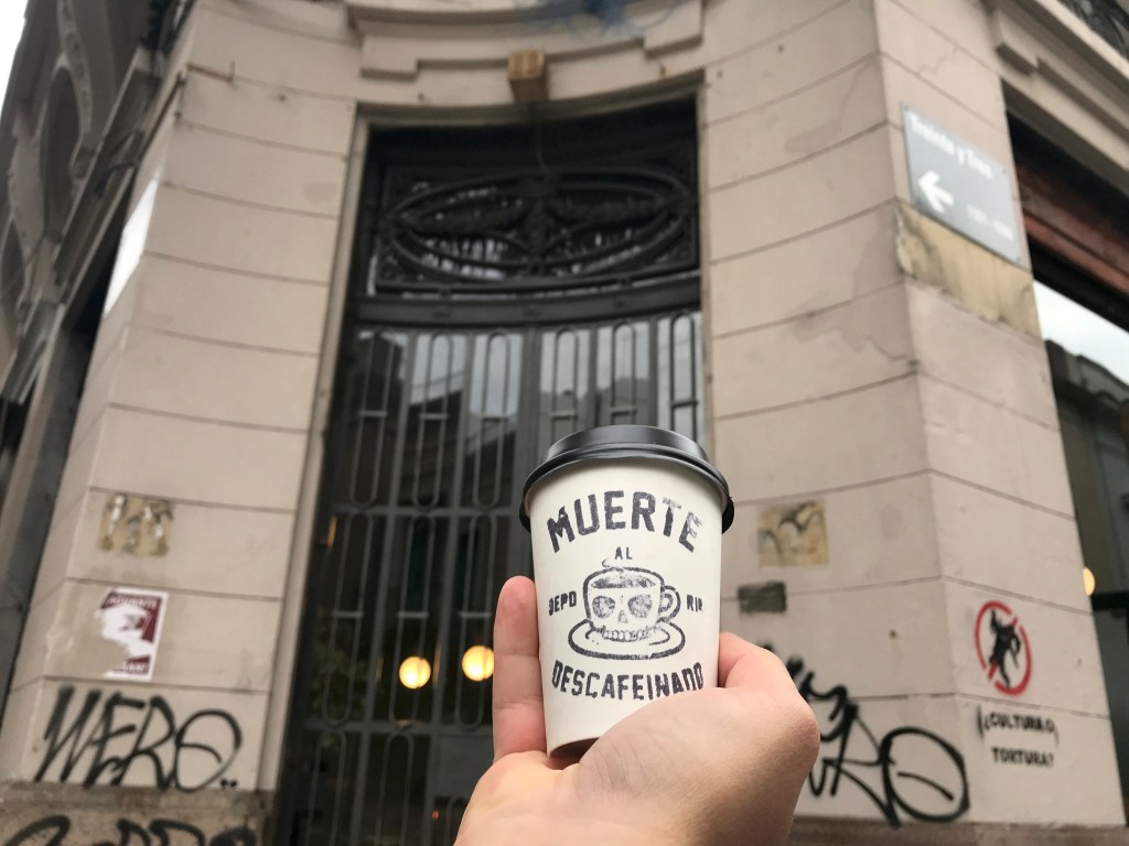 A photo of a cup of coffee being held near the front door to Nómade, a café in Montevideo, Uruguay. The photo is of the exterior / is an outside street view shot of the restaurant. Photo Courtesy of FoodWaterShoes