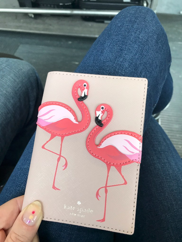 A photo of a neutral colored manicure created with Red Carpet Manicure gel polish in Camera Shy (natural), Losing My Vanity (purple), I Am So Honored (gold), Red Carpet Reddy (Red) and dotting pens. Also shown is a cute Kate Spade flamingo passport cover..